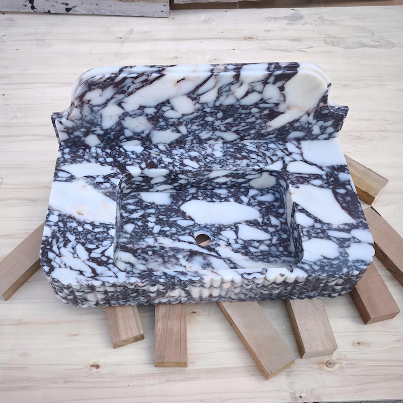 May include: A rectangular marble sink with a basin and a raised back. The sink features a white and dark purple veined pattern. The edges have a decorative, scalloped design. The sink is placed on wooden supports.