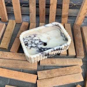 Op de afbeelding: Rechthoekige marmeren wastafel met een geschulpte rand. De wastafel heeft een witte en cr&egrave;mekleurige basis met donkergrijze adering. De wastafel staat tussen verschillende houten planken.