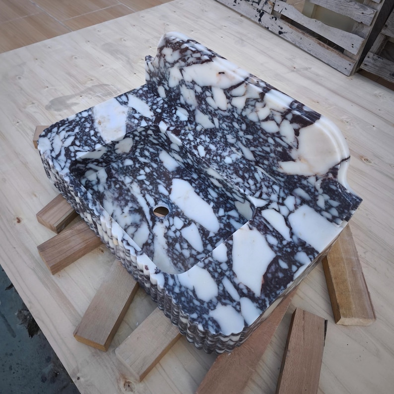 May include: A rectangular marble sink with a speckled pattern of white and dark gray. The sink has a curved front edge and a drain hole. It is resting on wooden supports.