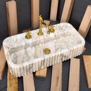 Op de afbeelding: Een rechthoekige marmeren wastafel met een gegroefde rand en gouden armaturen. De wastafel heeft een wit en beige geaderd patroon. De kraan en knoppen zijn goudkleurig. Verschillende houten planken omringen de wastafel.