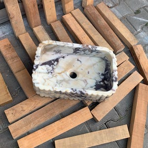 Op de afbeelding: Rechthoekige marmeren wastafel met een geschulpte rand. De wastafel heeft een cr&egrave;me- en bruin gemarmerd patroon en een centraal afvoergat. De wastafel is omgeven door verschillende houten planken.