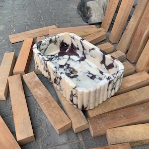 Op de afbeelding: Een rechthoekige marmeren wastafel met een gegroefde buitenkant. De wastafel heeft een witte basis met donker bordeauxrode en zwarte adering. De wastafel is omgeven door houten planken.