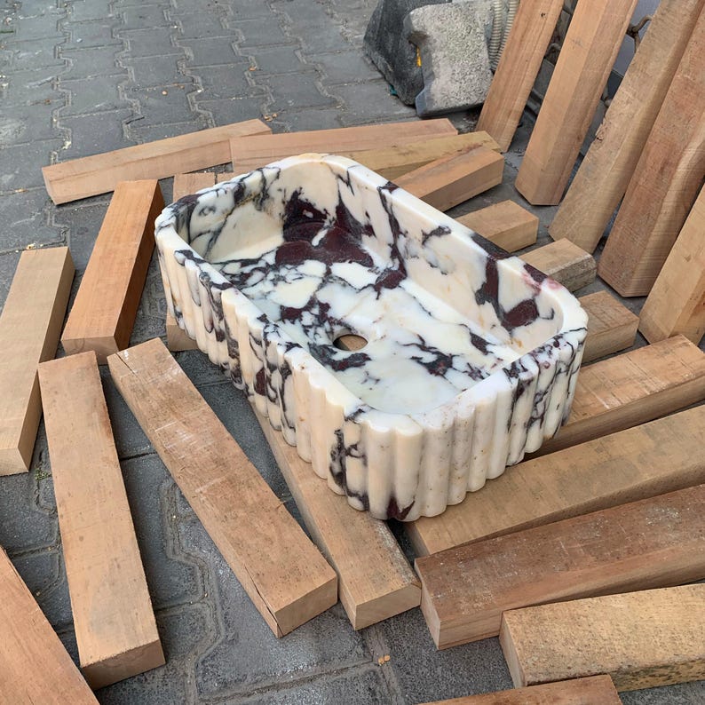 Op de afbeelding: Een rechthoekige marmeren wastafel met een gegroefde buitenkant. De wastafel heeft een witte basis met donker bordeauxrode en zwarte adering. De wastafel is omgeven door houten planken.