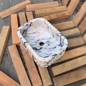 Op de afbeelding: Een rechthoekige, witte marmeren wastafel met donkergrijze adering. De wastafel heeft een geschulpte rand en een centraal afvoergat. Het is omgeven door houten planken.
