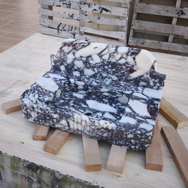 May include: A rectangular marble sink with a speckled pattern of white, black, and brown. The sink is supported by wooden blocks and has a curved back. The sink is a bathroom fixture.