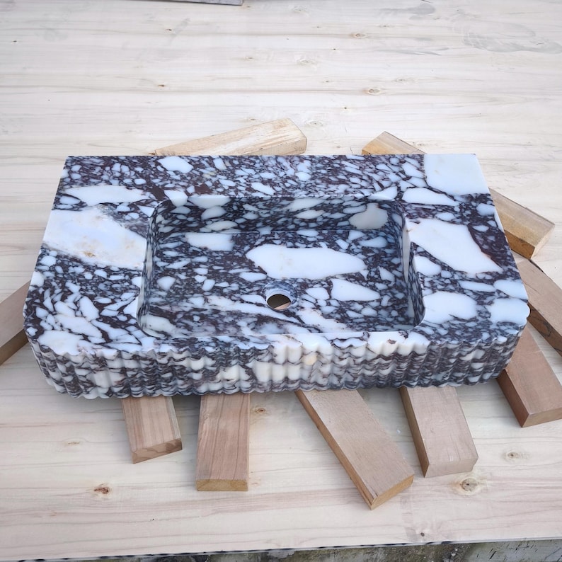 May include: A rectangular marble sink with a speckled pattern of white and dark gray. The sink has a basin with a drain hole and a decorative, textured edge. The sink is set on wooden supports.