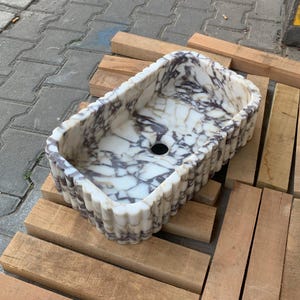 Op de afbeelding: Rechthoekige marmeren wastafel met een wit en paars geaderd patroon. De wastafel heeft een geschulpte rand en een centraal afvoergat. Hij rust op houten planken.