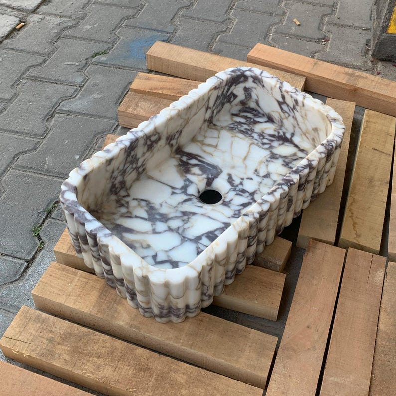 Op de afbeelding: Rechthoekige marmeren wastafel met een wit en paars geaderd patroon. De wastafel heeft een geschulpte rand en een centraal afvoergat. Hij rust op houten planken.