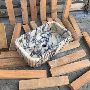 Op de afbeelding: Een rechthoekige marmeren wastafel met een gegroefde buitenkant. De wastafel heeft een wit en grijs gemarmerd patroon en een centraal afvoergat. De wastafel is omgeven door houten planken.
