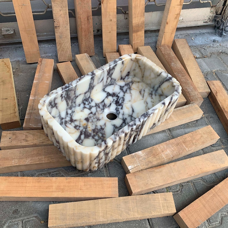 Op de afbeelding: Een rechthoekige marmeren wastafel met een gegroefde buitenkant. De wastafel heeft een wit en grijs gemarmerd patroon en een centraal afvoergat. De wastafel is omgeven door houten planken.