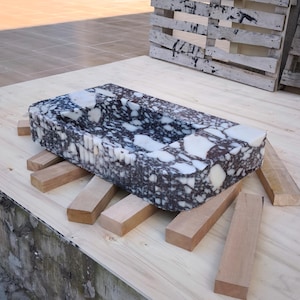 May include: A rectangular stone sink with a speckled black and white pattern. The sink is supported by several wooden beams and rests on a light-colored wooden surface. The sink's interior is a smooth, curved basin.