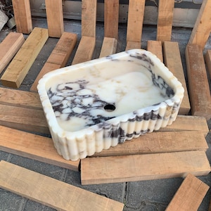 Op de afbeelding: Rechthoekige marmeren wastafel met een cr&egrave;me- en zwart patroon. De wastafel heeft een gegolfde rand en een centraal afvoergat. De wastafel wordt gepresenteerd op een oppervlak van houten planken.
