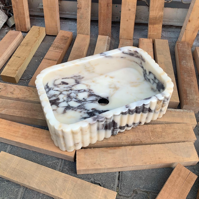 Op de afbeelding: Rechthoekige marmeren wastafel met een cr&egrave;me- en zwart patroon. De wastafel heeft een gegolfde rand en een centraal afvoergat. De wastafel wordt gepresenteerd op een oppervlak van houten planken.