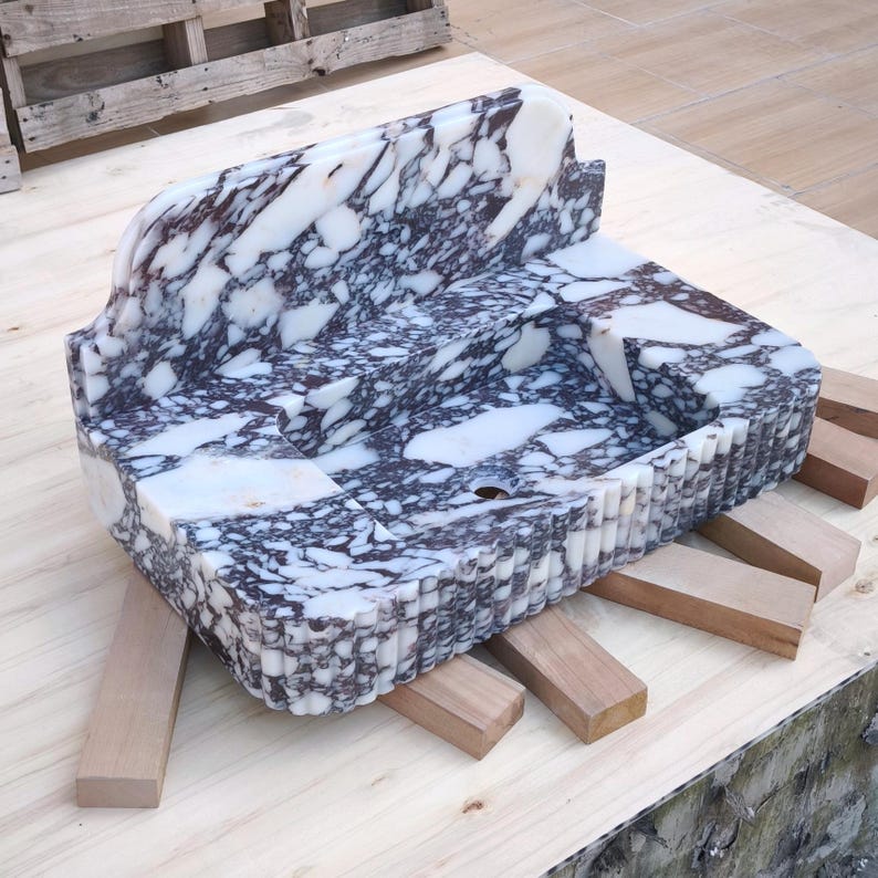 May include: A rectangular marble sink with a built-in backsplash. The sink features a white and dark purple marbled pattern. The front edge has a carved, ridged design. The sink is supported by wooden blocks on a wooden surface.