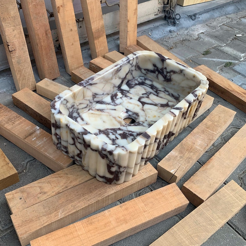 Op de afbeelding: Een rechthoekige marmeren wastafel met een cr&egrave;me- en donkerpaars geaderd patroon. De wastafel heeft een gegroefd exterieurontwerp en een centraal afvoergat. De wastafel wordt op houten planken gepresenteerd.