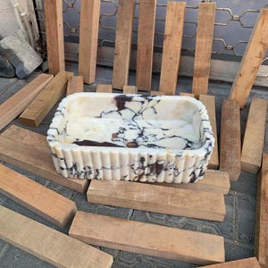 Op de afbeelding: Rechthoekige marmeren wastafel met een cr&egrave;me- en bruinpatroon. De wastafel heeft een gegroefde buitenkant en een centraal afvoergat. De wastafel is omgeven door houten planken.