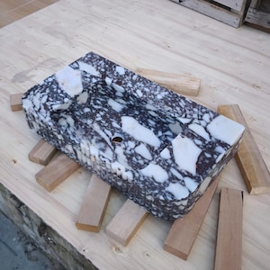 May include: A rectangular marble sink with a speckled pattern of white and dark purple. The sink has a rounded interior basin and a single drain hole. It is displayed on a light wooden surface with several wooden supports.
