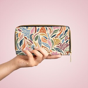 May include: A rectangular wallet with a colorful floral pattern. The wallet features a cream background with a repeating design of leaves and flowers in shades of blue, orange, pink, and green. It has a gold zipper and is held by a hand.