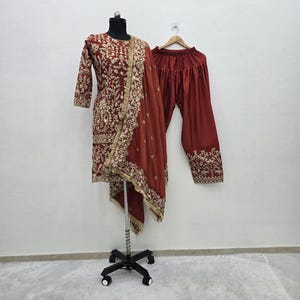 Könnte beinhalten: Ein maroonfarbenes, besticktes Salwar-Kameez-Set. Das Set umfasst eine langärmlige Tunika mit aufwendiger Goldstickerei, eine passende Hose mit weitem Bein und einen drapierten Schal. Das Outfit wird auf einer Schaufensterpuppe und einem Kleiderbügel präsentiert.