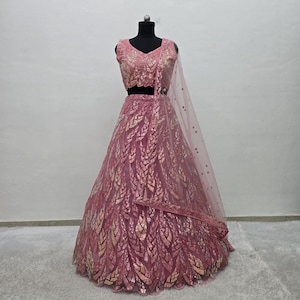 May include: A pink lehenga set on a black mannequin. The set includes a sleeveless top and a long skirt, both adorned with gold leaf-like patterns. A sheer, dotted dupatta drapes over the shoulder. The outfit is designed for formal occasions.