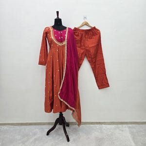 May include: A rust-colored long-sleeved dress with a magenta bodice featuring gold embroidery and buttons, paired with matching rust-colored trousers and a long magenta dupatta with a gold-trimmed edge, displayed on a black mannequin.