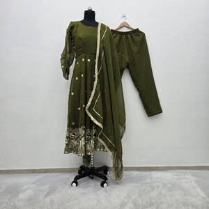 May include: An olive green Indian suit set, including a long-sleeved tunic with floral embroidery and a matching dupatta. The tunic is displayed on a mannequin, with matching pants hanging on a wooden hanger. The dupatta has a gold trim.