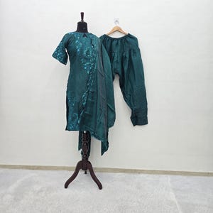 May include: A dark teal three-piece outfit consisting of a knee-length tunic with intricate embroidery around the neckline and sleeves, a matching draped shawl, and pleated trousers. The ensemble is displayed on a mannequin and a wooden hanger.