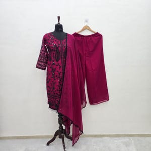 Puede incluir: Un conjunto de tres piezas de color magenta, compuesto por una túnica con bordados negros, pantalones de pernera ancha y una bufanda transparente, expuesto sobre un maniquí y una percha. El conjunto se encuentra sobre un fondo blanco liso. La túnica tiene adornos.
