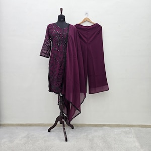 以下が含まれることがあります： マネキンとハンガーに展示された、プラム色の3ピースの衣装。 黒いレースのディテールが施された長袖トップス、お揃いのワイドレッグパンツ、シアーなスカーフが含まれています。 衣装は無地の白い背景に設定されています。