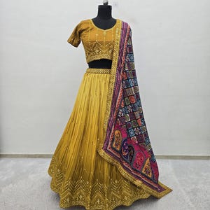 May include: A mustard yellow lehenga choli set featuring a short-sleeved blouse and a pleated floor-length skirt, both adorned with intricate gold embroidery and small mirrors. A multicolored dupatta with a geometric and floral print is draped.
