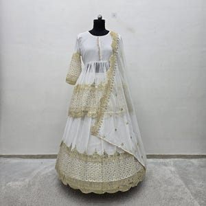May include: A white three-piece ensemble featuring a peplum-style top with three-quarter sleeves, a long flared skirt, and a matching dupatta. The garments are adorned with intricate gold embroidery, metallic sequins, and scalloped hem details.