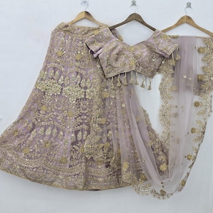 May include: A lavender lehenga set featuring a floor-length skirt, a short-sleeved blouse, and a sheer dupatta. The ensemble is heavily embellished with intricate gold embroidery, sequins, and metallic thread work throughout the fabric.