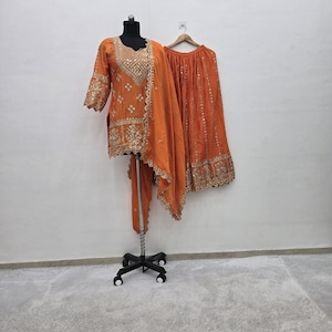 Könnte beinhalten: Ein orangefarbenes, besticktes Salwar-Kameez-Set, das auf einer Schaufensterpuppe und einem Kleiderbügel präsentiert wird. Das Set umfasst eine Tunika, eine Hose und einen Schal, alle mit goldener Stickerei verziert. Das Outfit ist aus fließendem Stoff gefertigt.