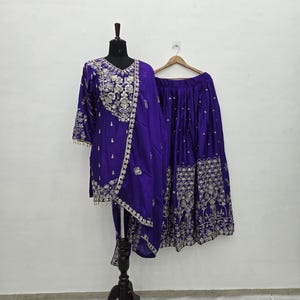 May include: A vibrant purple ensemble featuring a tunic, skirt, and scarf, all adorned with intricate gold embroidery. The tunic is displayed on a mannequin, while the skirt hangs on a wooden hanger. The outfit is designed with a traditional aesthetic.