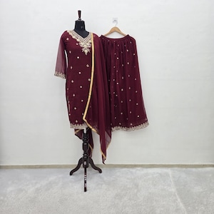 Pode incluir: Um conjunto bordô com uma túnica de mangas compridas com bordados dourados e uma saia a condizer. A túnica é exibida num manequim e a saia está pendurada num cabide de madeira. O traje é adornado com detalhes dourados e pequenos enfeites.