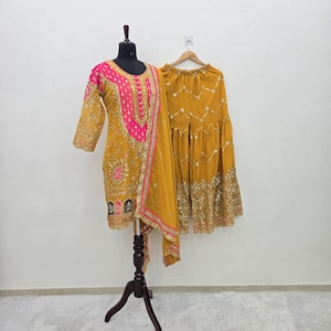 May include: Mustard yellow embroidered salwar kameez set with a pink and gold embellished neckline and sleeves. The set includes a matching dupatta and flared pants, displayed on a mannequin and hanger.