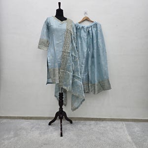 May include: A light blue three-piece outfit consisting of a long-sleeved tunic, a wide-leg skirt, and a matching dupatta. The garments feature silver metallic embroidery and sequin detailing along the hemlines and throughout the fabric.