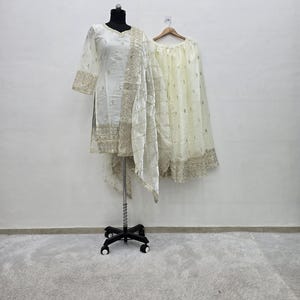 Könnte beinhalten: Ein cremefarbenes, besticktes dreiteiliges Outfit, das auf einer Schaufensterpuppe und einem Kleiderbügel präsentiert wird. Das Ensemble umfasst eine Tunika, einen passenden Schal und einen Rock. Die Tunika und der Rock weisen aufwendige goldene Stickereien auf.