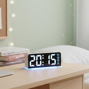 May include: A digital alarm clock displaying 20:15 with a blue LED light. The clock sits on a wooden desk next to a laptop, books, and a small plant. A corkboard with photos and fairy lights is in the background.