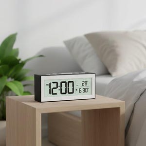 May include: A black and silver digital alarm clock displaying the time 12:00, with the date and alarm time visible. The clock sits on a light brown wooden nightstand next to a bed with white bedding. A potted plant is in the background.