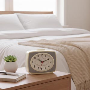Puede incluir: Un despertador de estilo retro con esfera gris y marco crema está sobre una mesita de noche de madera. El reloj marca las 10:10. Una pequeña suculenta en maceta blanca, un cuaderno y un bolígrafo también están sobre la mesita. Al fondo, una cama con ropa de cama blanca.