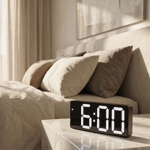 May include: A black digital alarm clock displaying 6:00 with white LED numerals. The clock sits on a marble bedside table next to a bed with beige bedding and pillows. The room has neutral tones and natural light.
