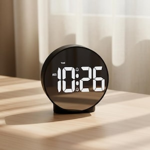 Op de afbeelding: Een zwarte digitale wekker met de tekst "10:26 AM DIN" in het wit op een spiegeloppervlak. De klok heeft een ronde wijzerplaat en een rechthoekige basis, geplaatst op een licht houten oppervlak.