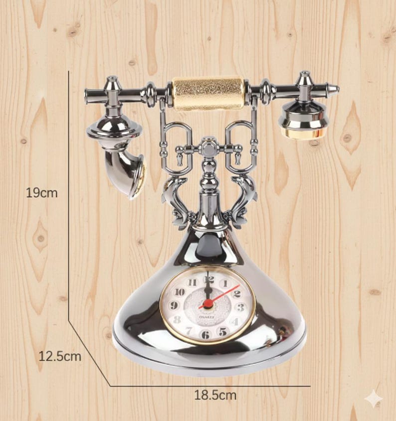 Może przedstawiać: Srebrny, zabytkowy telefon z tarczą zegara. Telefon ma 19 cm wysokości, 12,5 cm szerokości i 18,5 cm długości. Posiada złoty uchwyt i zegar z rzymskimi cyframi. Telefon stoi na drewnianej powierzchni.