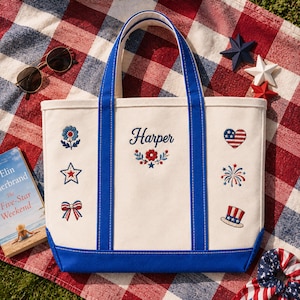 Könnte beinhalten: Eine cremefarbene Canvas-Tragetasche mit blauen Akzenten und Griffen, bestickt mit patriotischen Symbolen und dem Namen "Harper". Die Tasche hat ein rot-weiß-blaues Farbschema, ideal für einen Sommerausflug.