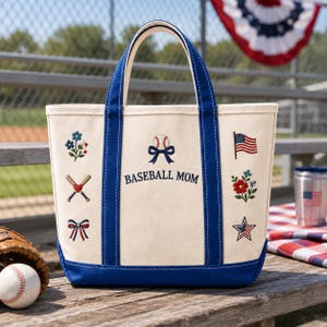 May include: A cream-coloured canvas tote bag with blue trim and handles. The front features embroidered designs including a baseball with a blue bow, the text BASEBALL MOM, a small American flag, a star, flowers, and crossed baseball bats.