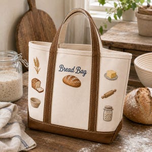 May include: A cream-colored canvas tote bag with brown trim and handles. The bag features the text Bread Bag in blue script, surrounded by embroidered illustrations of wheat, a loaf of bread, a proofing basket, butter, a rolling pin, and a jar.