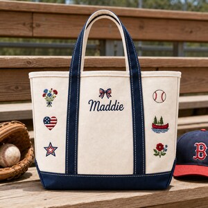 May include: A cream-coloured canvas tote bag with navy blue handles and base. The bag features embroidered designs including a bouquet, a heart with a flag pattern, a star, a baseball, a canoe with trees, and a flower. The name Maddie is embroidered in navy blue script below a small bow.