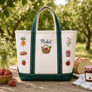 May include: A white canvas tote bag with dark green trim and handles. The bag features embroidered designs including carrots, a loaf of bread, a strawberry, a flower bouquet, a jar of jam, an apple, and a basket with the name Mabel embroidered above.