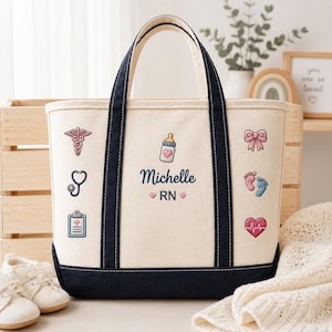 May include: Cream-colored tote bag with navy blue accents and handles. The bag features embroidered medical and baby-themed icons, the name "Michelle", and "RN". A stethoscope, baby bottle, and heart are among the designs.